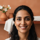 Close-up portrait of a smiling South Asian woman, looking at the camera with a happy expression. Her face is well-lit, and th